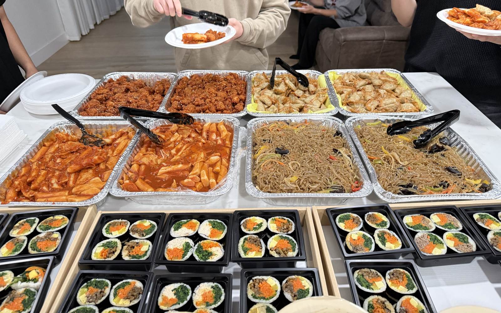 Trays of sweet chili chicken, tteokbokki, japchae and mandu with kimbap boxes at a Minari catered lunch